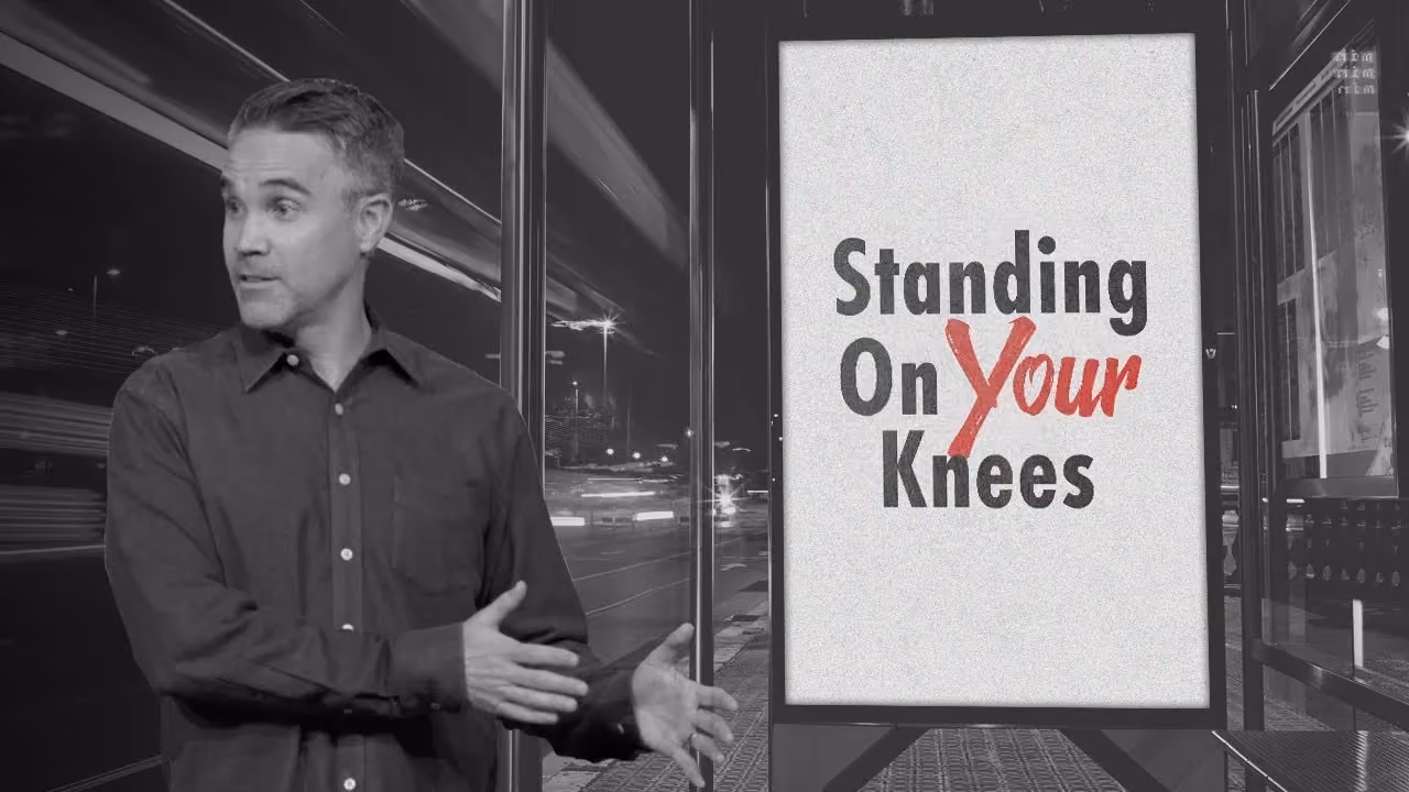 Man gesturing beside sign reading Standing On Your Knees with 'Your' crossed out in red.