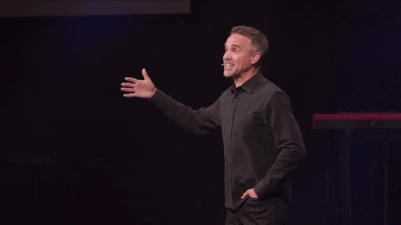 Man gesturing while speaking on a dark stage with a keyboard in background.