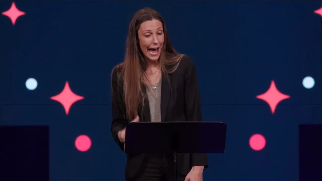 Woman speaking at podium with colorful star and circle graphics on dark blue background.