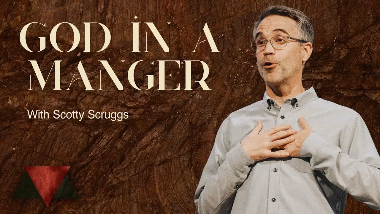 God in a Manger with Scotty Scruggs, hands to his chest against a dark wood backdrop.