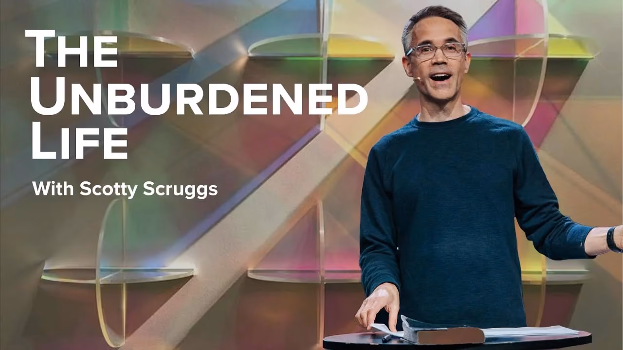 The Unburdened Life with Scotty Scruggs, at a lectern before panels of coloured glass.