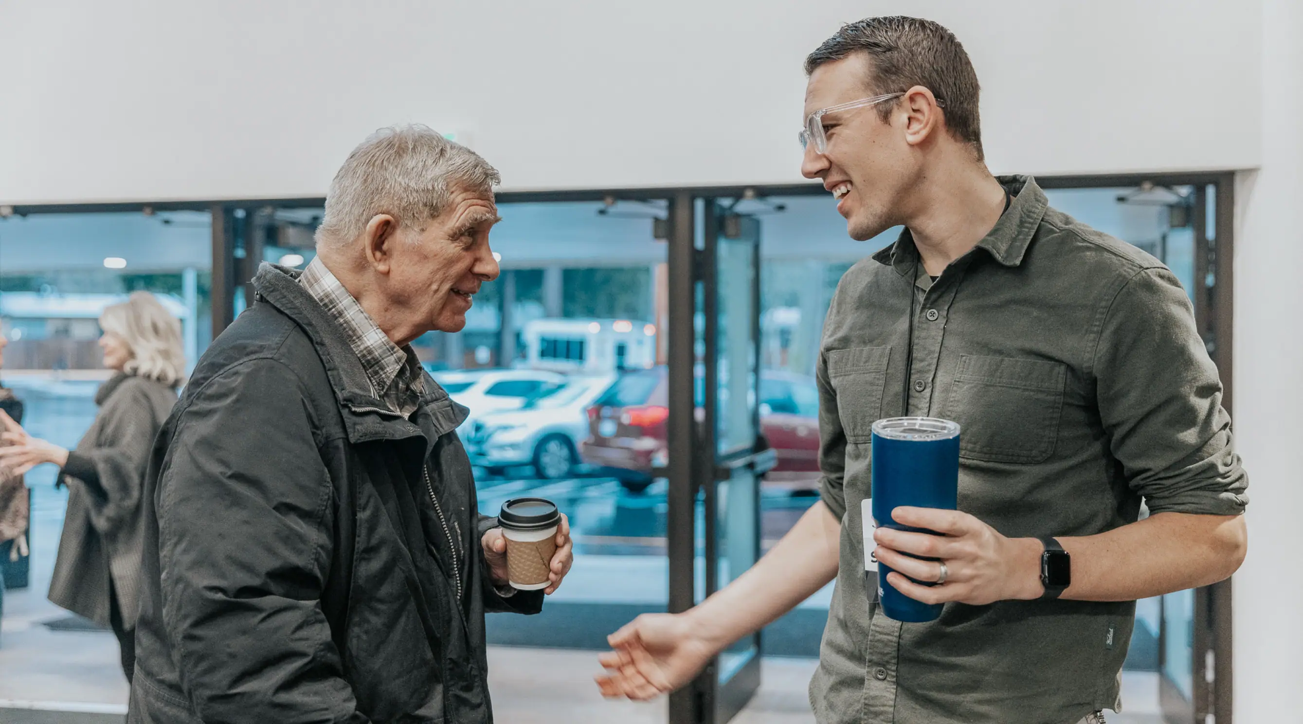 Photo of an older man and a younger man smiling and chatting outside a building, each holding a drink