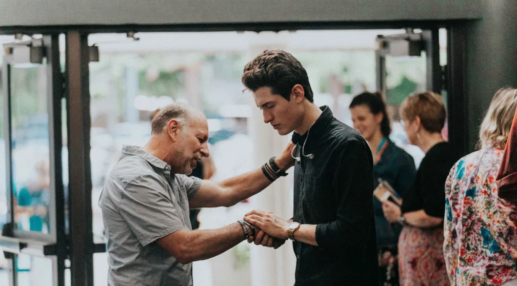 A man holding a young man's hands in a moment of prayer.