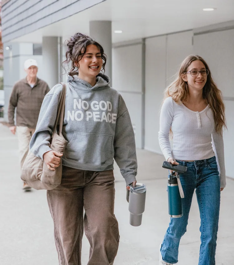 Photo of two smiling young women walking together indoors, one wearing a hoodie reading 'NO GOD NO PEACE'