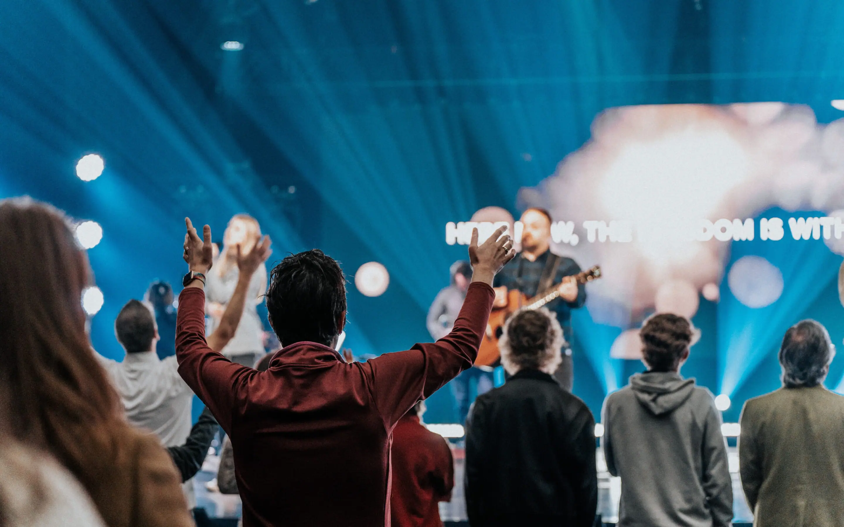 Photo from behind the congregation of people raising their hands toward the stage during a worship service, with song lyrics displayed on a screen