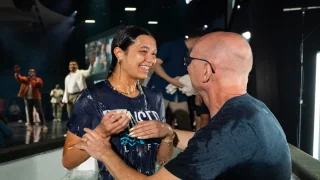 Photo of a smiling woman being baptized in a pool by a man, with onlookers in the background