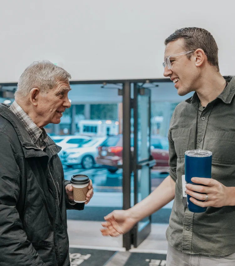 Photo of an older man and a younger man smiling and chatting outside a building, each holding a drink
