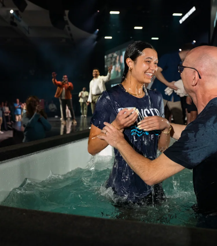 Photo of a young woman smiling as a man baptizes her in a baptism pool, with onlookers celebrating in the background