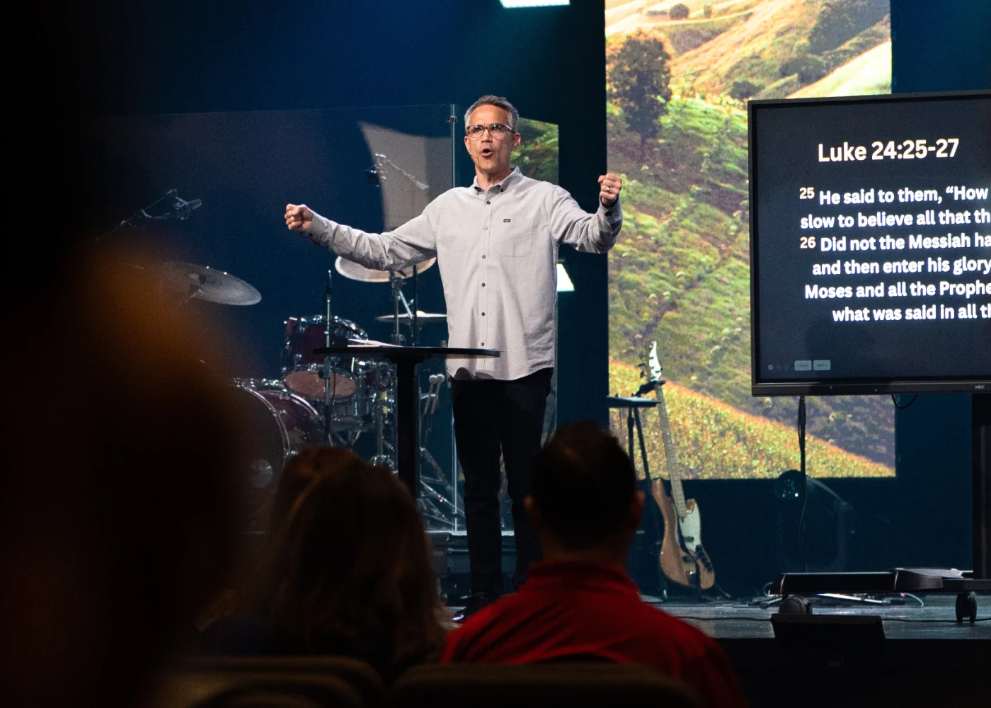 Photo of a pastor preaching on stage with arms raised, a Bible passage displayed on the screen behind him