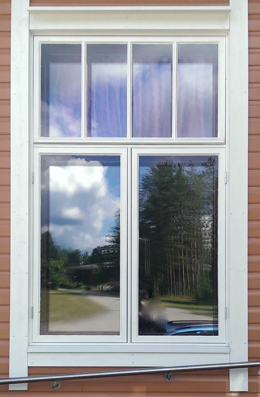 Valkoinen perinneikkuna, jonka lasipinnoissa heijastuu pilvinen taivas ja mäntymetsä.