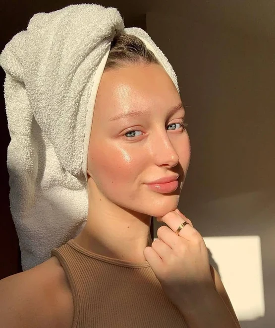 Young woman with clear skin and a towel wrapped around her head, posing in warm sunlight.