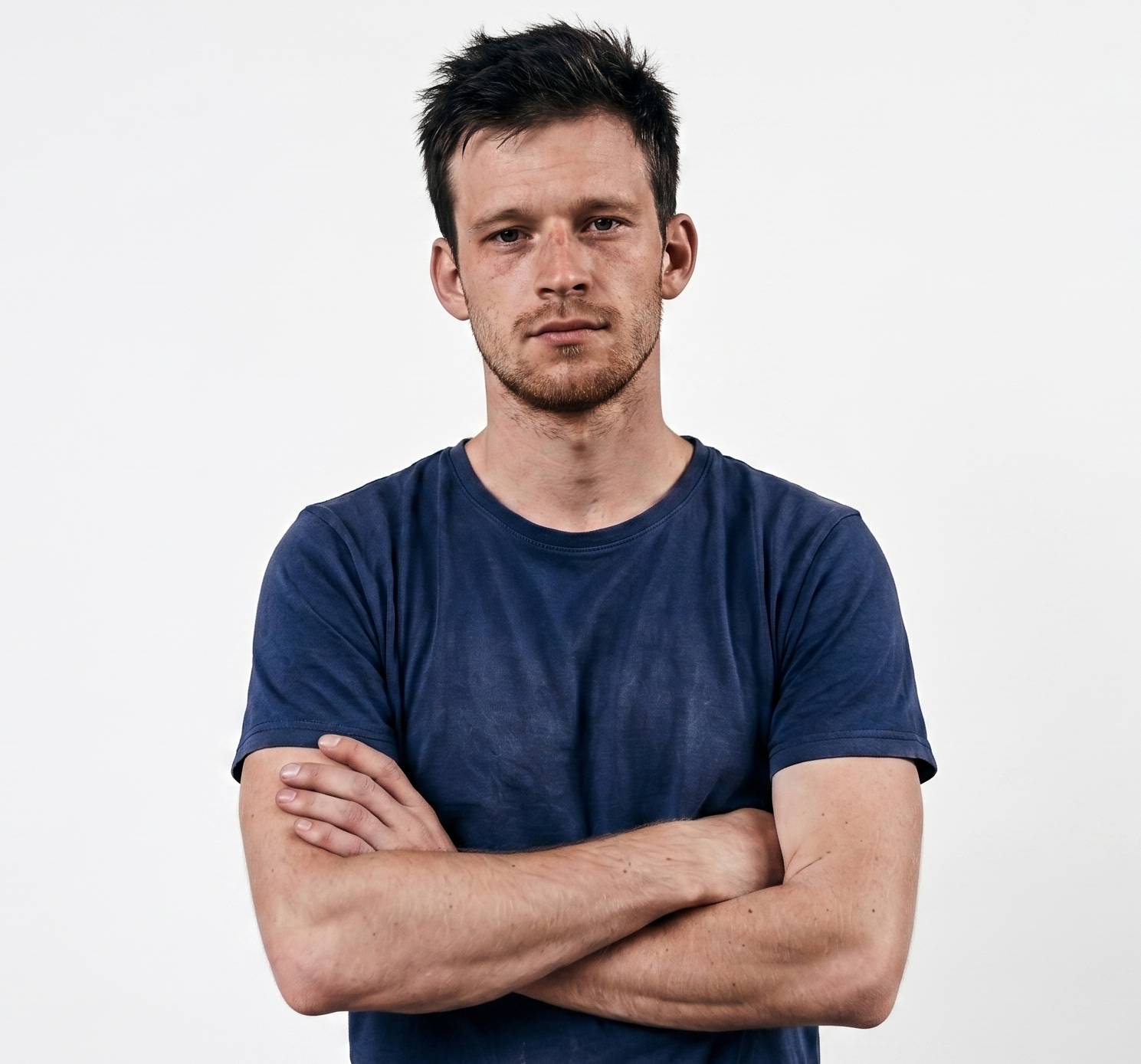 Serious young man with short dark hair and stubble wearing a navy blue t-shirt with arms crossed.