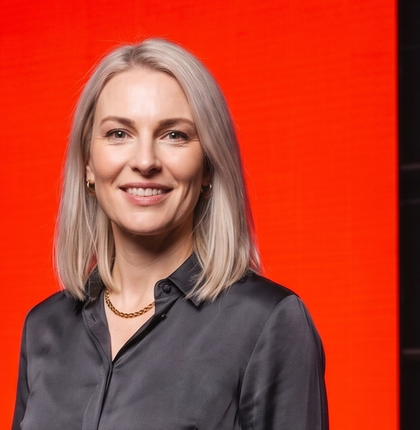Smiling woman with blonde hair wearing a black blouse and gold necklace against a red background.