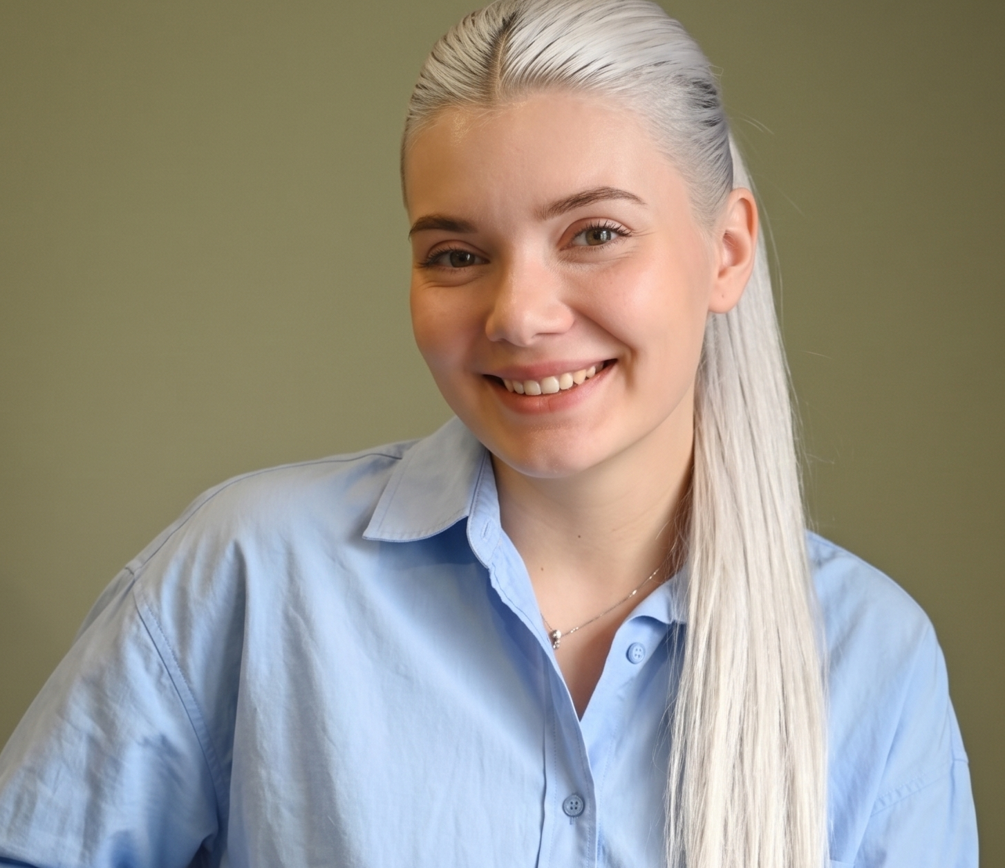 Young woman with long silver hair smiling, wearing a light blue buttoned shirt against a plain background.