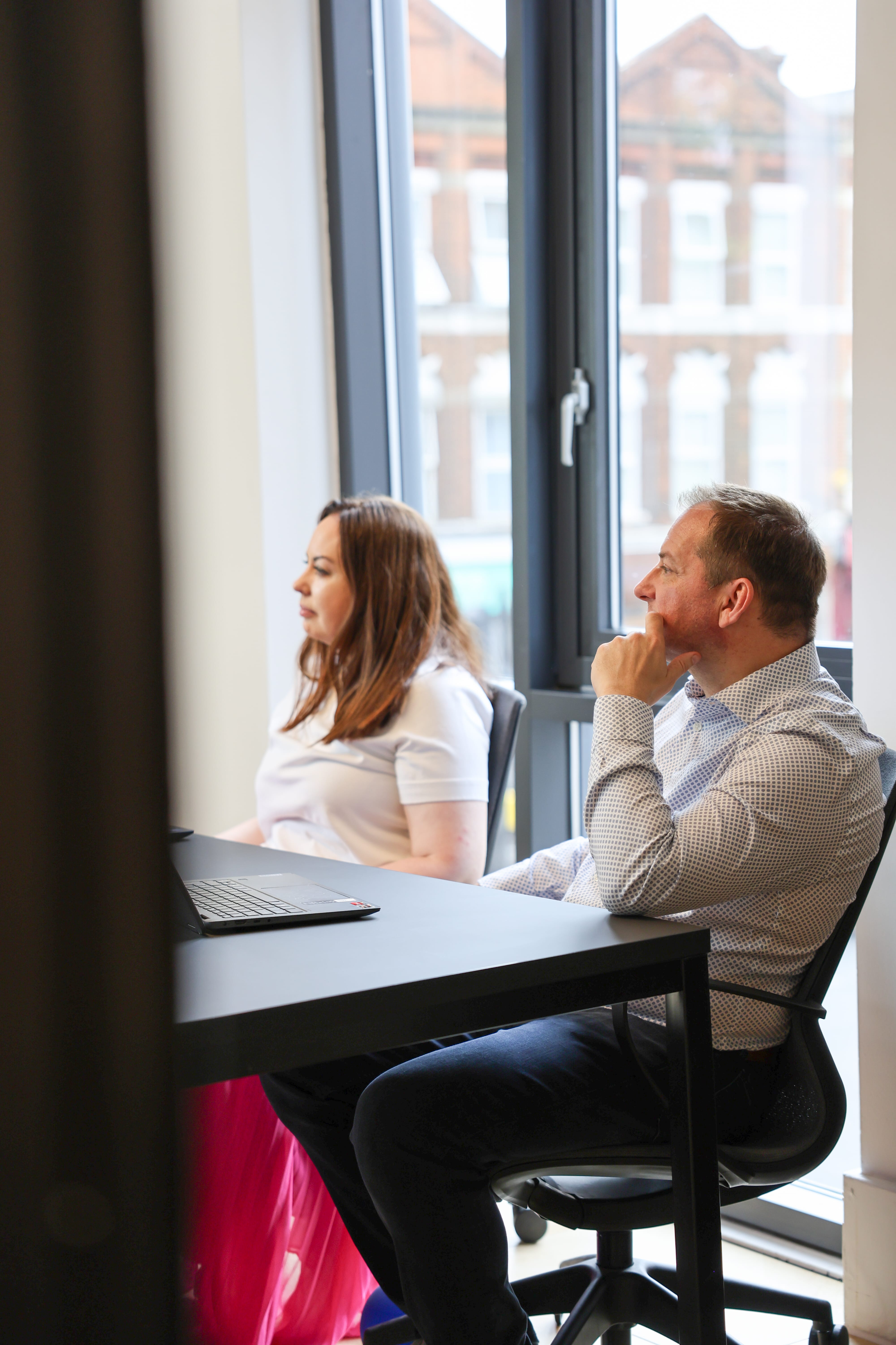 A man and a woman sitting side by side at a desk with a laptop, looking ahead thoughtfully in a bright room with large windows showing building exteriors outside.