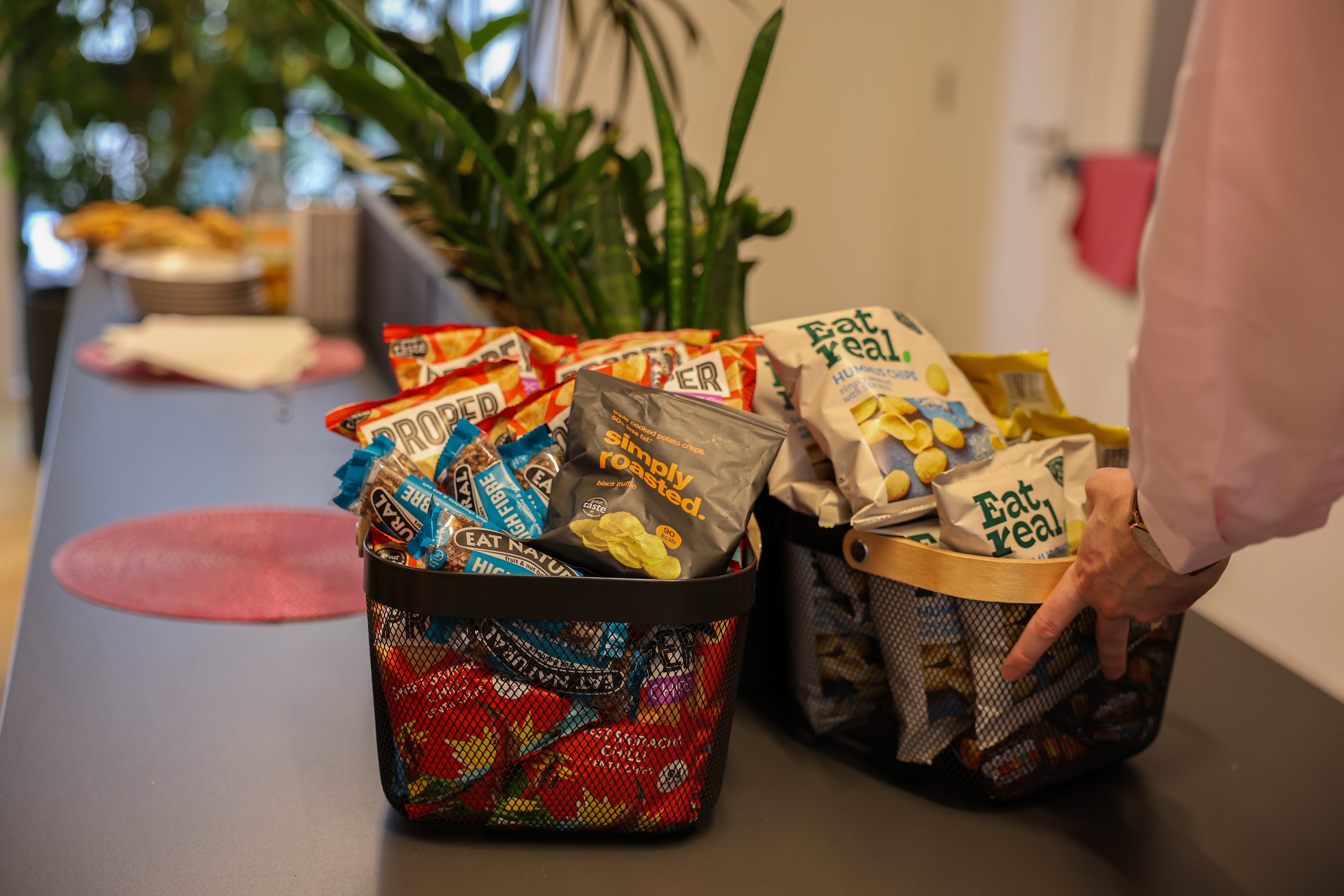 Two black mesh baskets filled with various snack bags including Simply Roasted potato crisps, Eat Real hummus chips, and other snack brands on a counter, with a person's hand reaching for one basket.