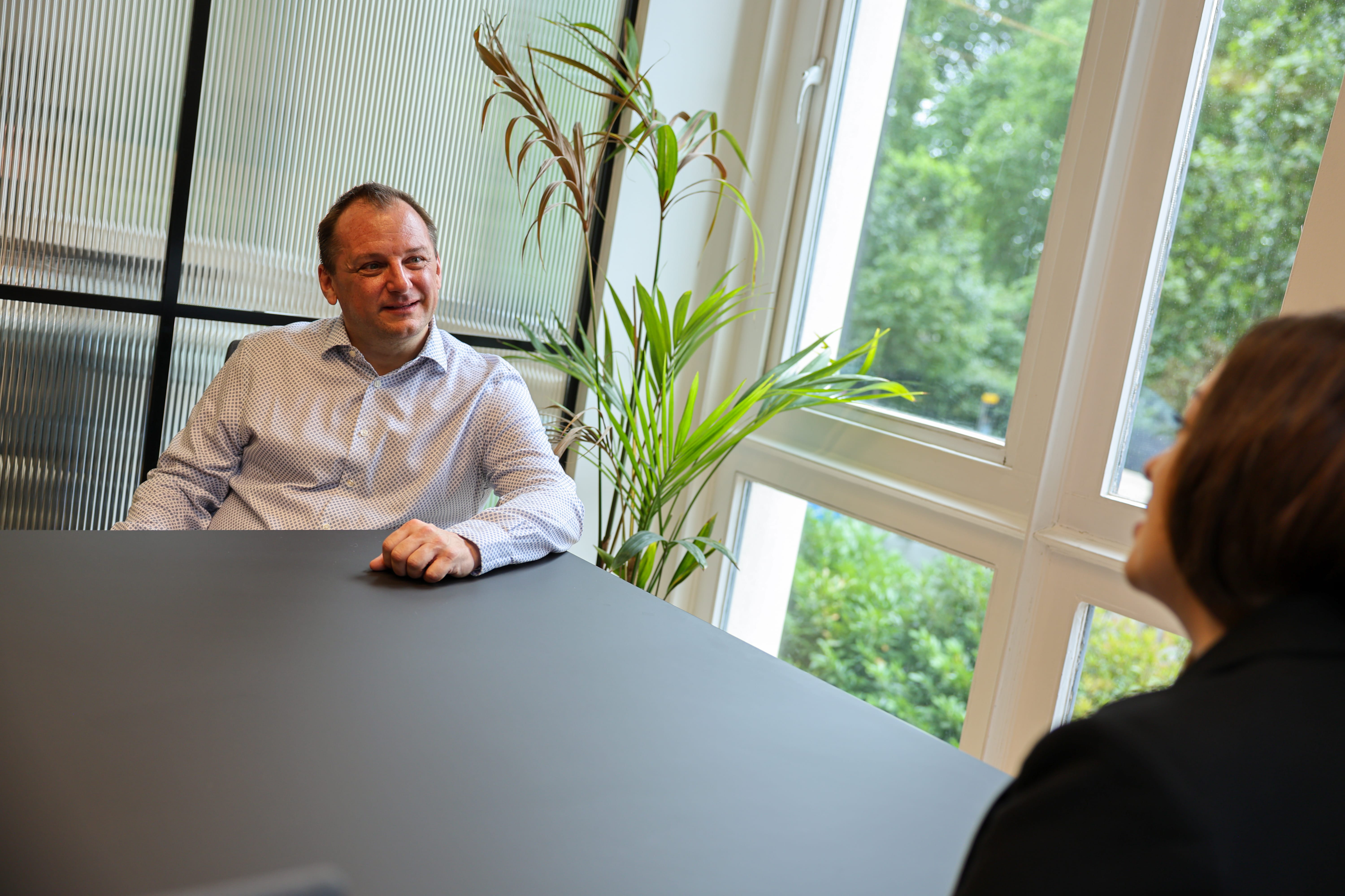 Man in a white patterned shirt sitting at a black table, engaging in conversation with a woman in a black jacket, near a window with green plants visible outside and indoor plants beside them.