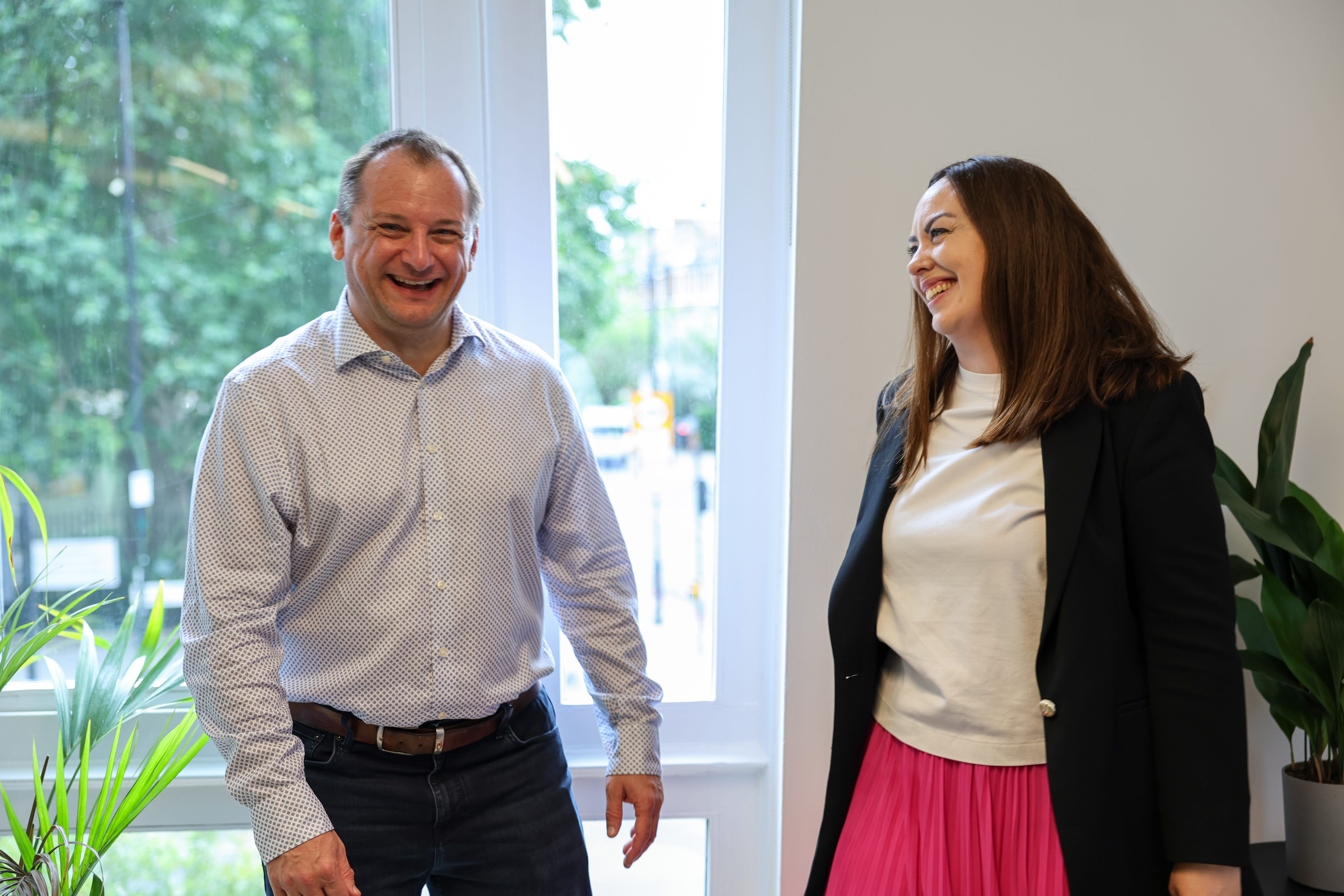 A man and a woman standing indoors near a window with green plants on either side, both smiling and looking at each other.