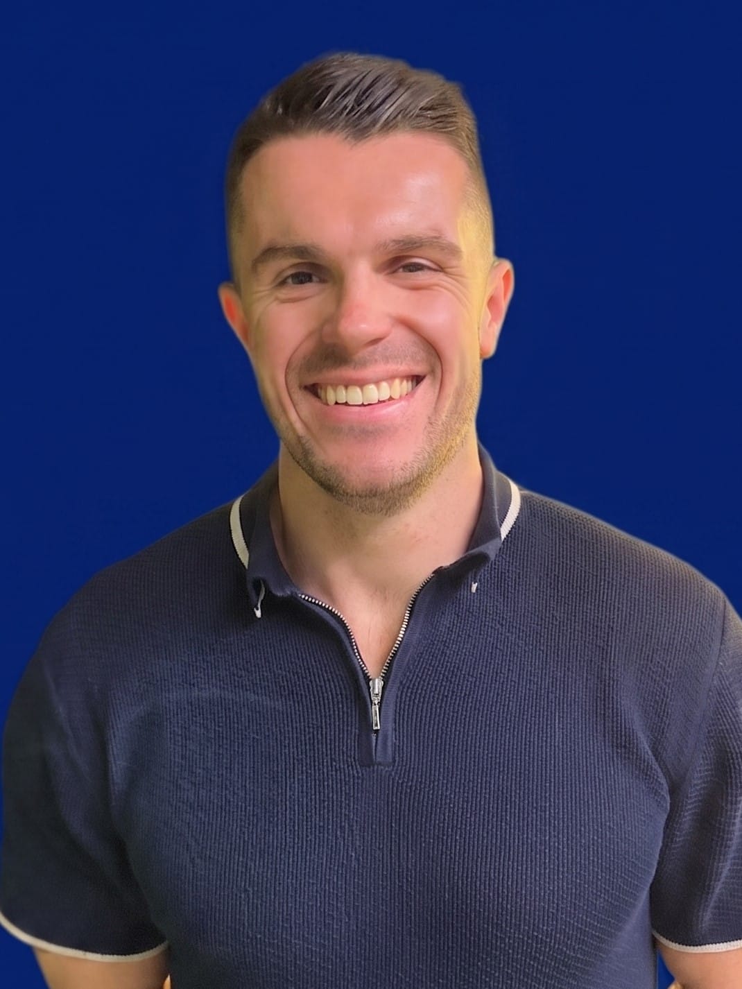 Smiling man with short light brown hair wearing a navy blue quarter-zip sweater against a solid blue background.