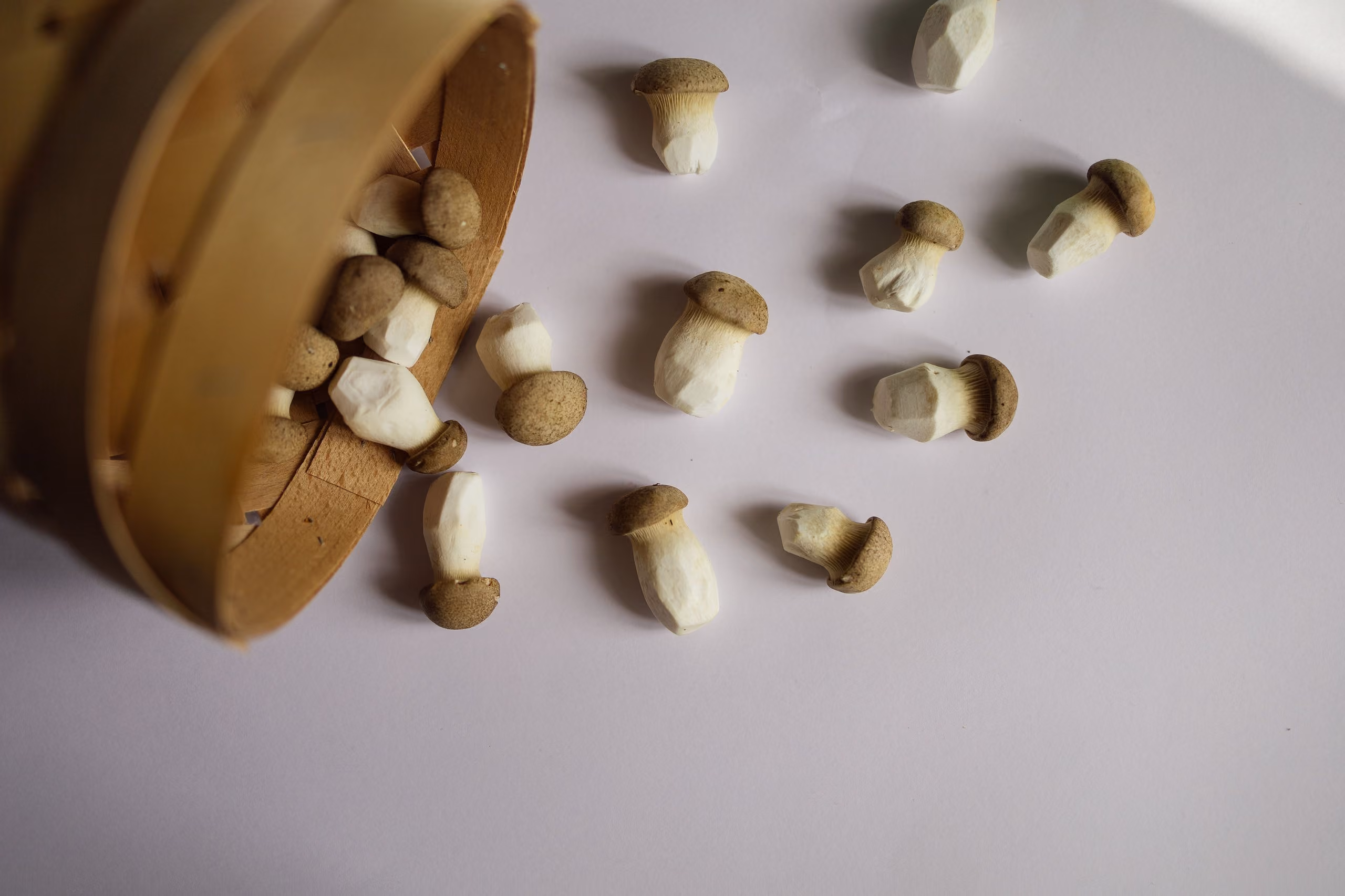 Small brown mushrooms spilling from a tipped wooden basket onto a light surface.