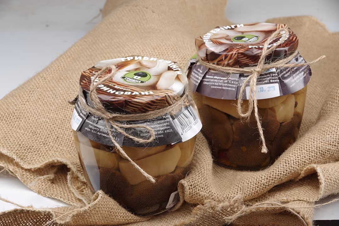 Two glass jars filled with pickled mushrooms wrapped in brown fabric and tied with twine.