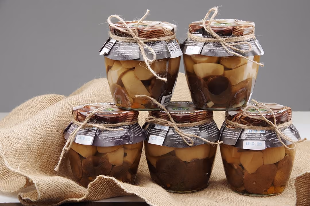 Five jars of preserved mushrooms stacked on burlap fabric with twine tied around lids.