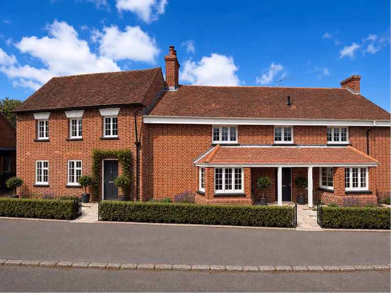 Red brick two-story house under blue sky with white clouds and trimmed bushes in front.