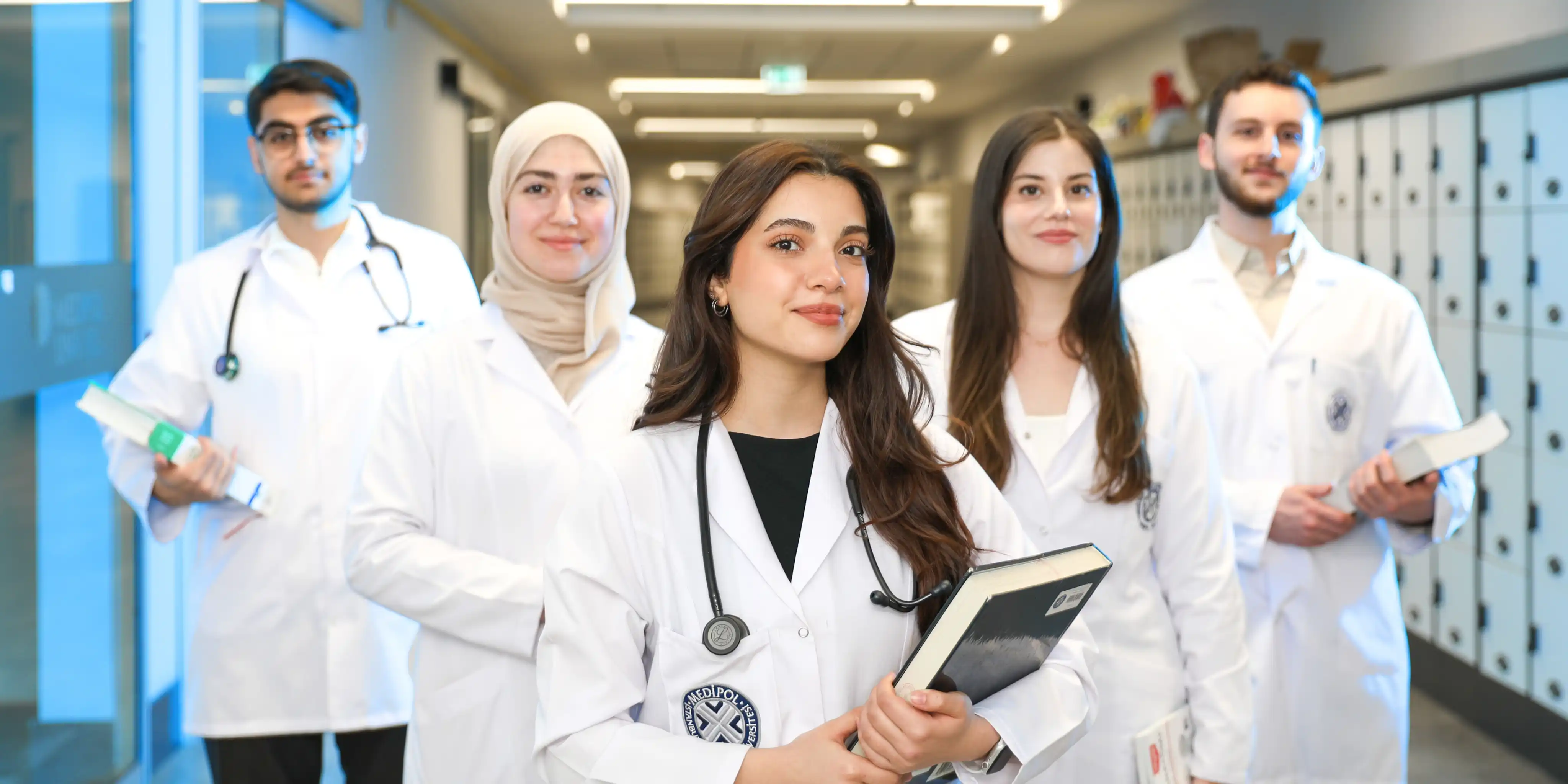 Five medical students in white coats standing in a hallway, holding books and wearing stethoscopes.