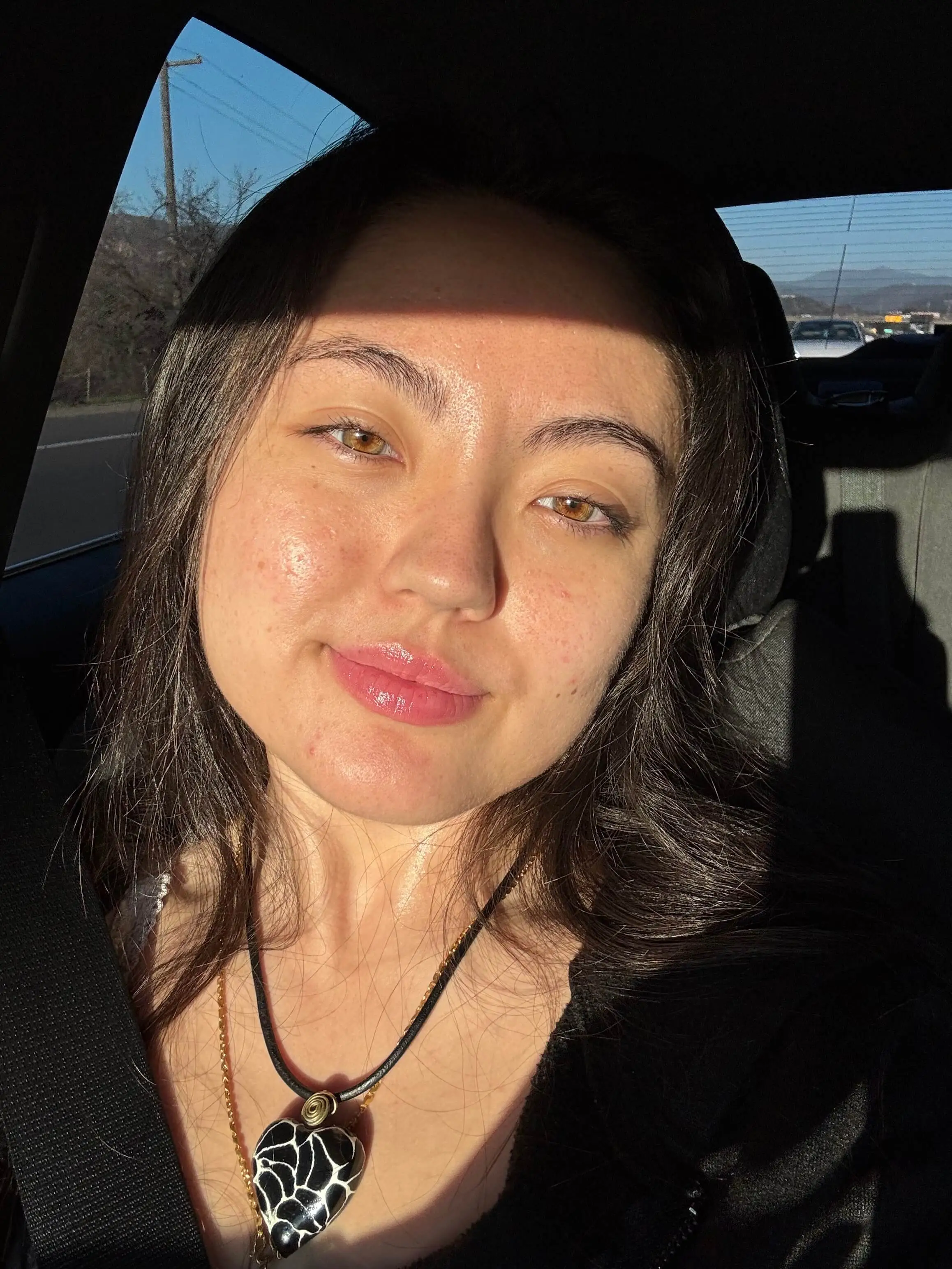 Alexisn. a young woman with light brown eyes and glossy lips sitting in a car wearing a black top and seatbelt, with sunlight casting a shadow across her forehead and a black and white heart-shaped pendant necklace visible.