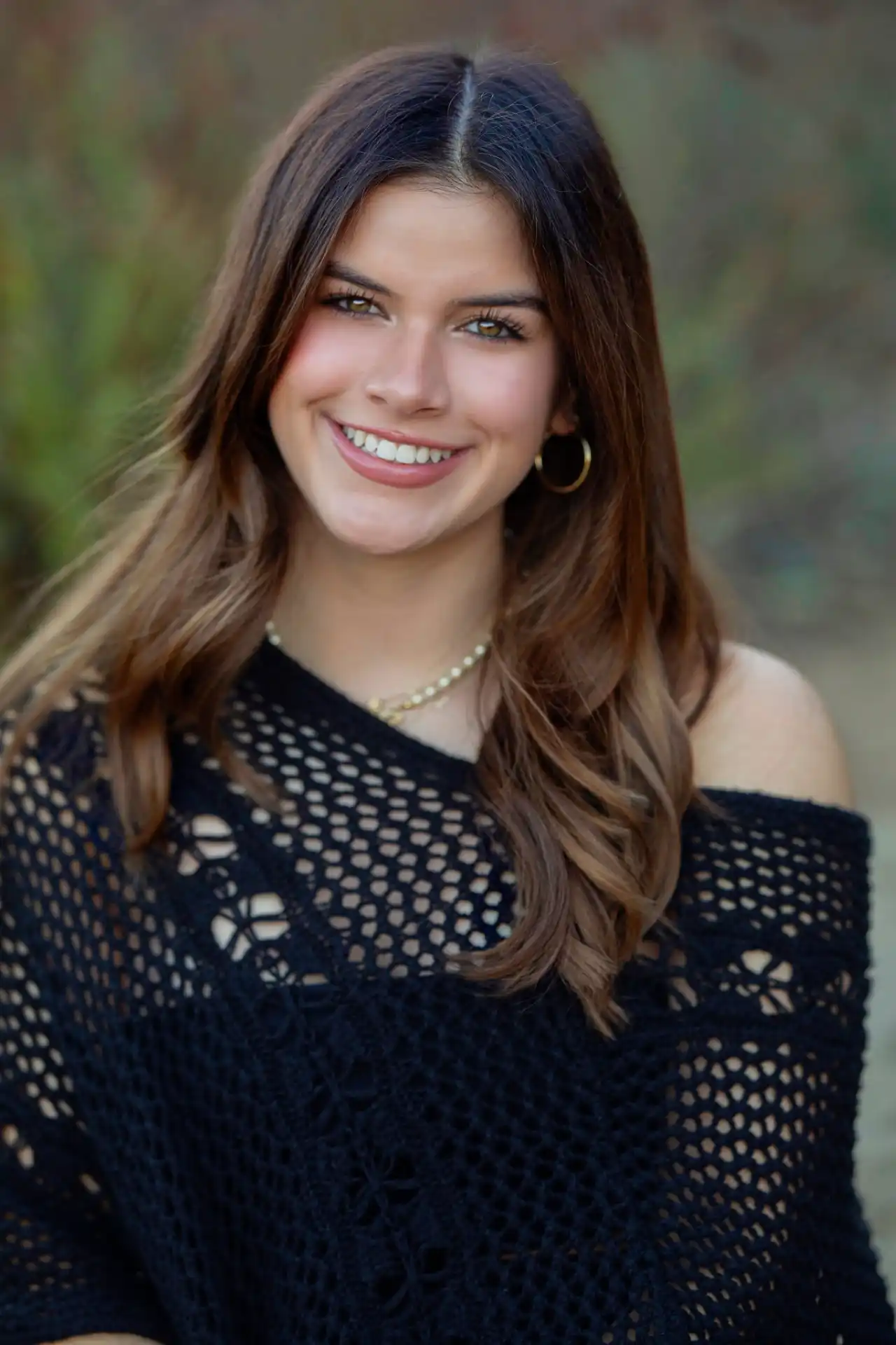 Anna, an smiling young woman with long brown hair wearing a black knitted off-shoulder top, gold hoop earrings, and a pearl necklace, standing outdoors with a blurred natural background.