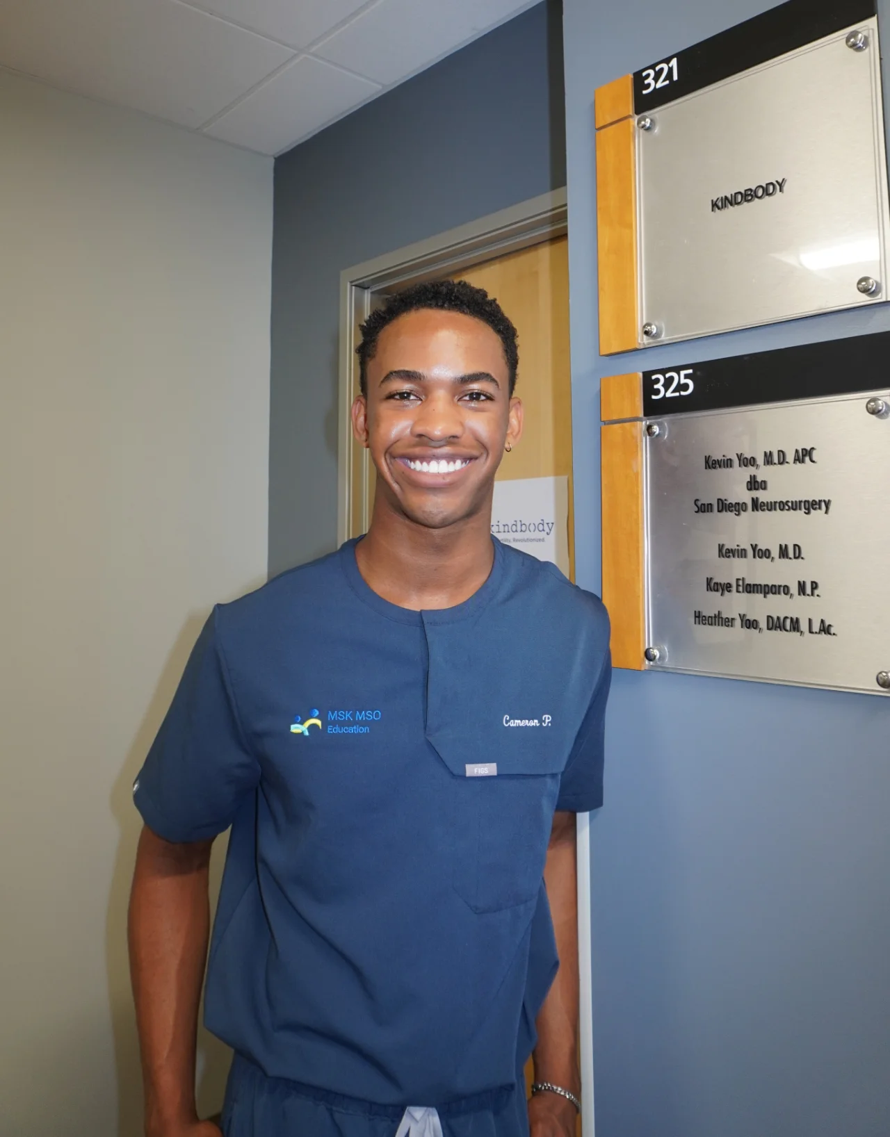 Cameron, a smiling young man in blue medical scrubs standing in a hallway next to office signs for Kindbody and San Diego Neurosurgery.