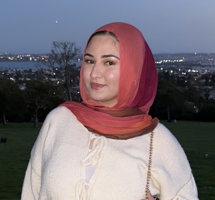 Nadia, a woman wearing a gradient red and orange headscarf and a white sweater standing outdoors with a cityscape.