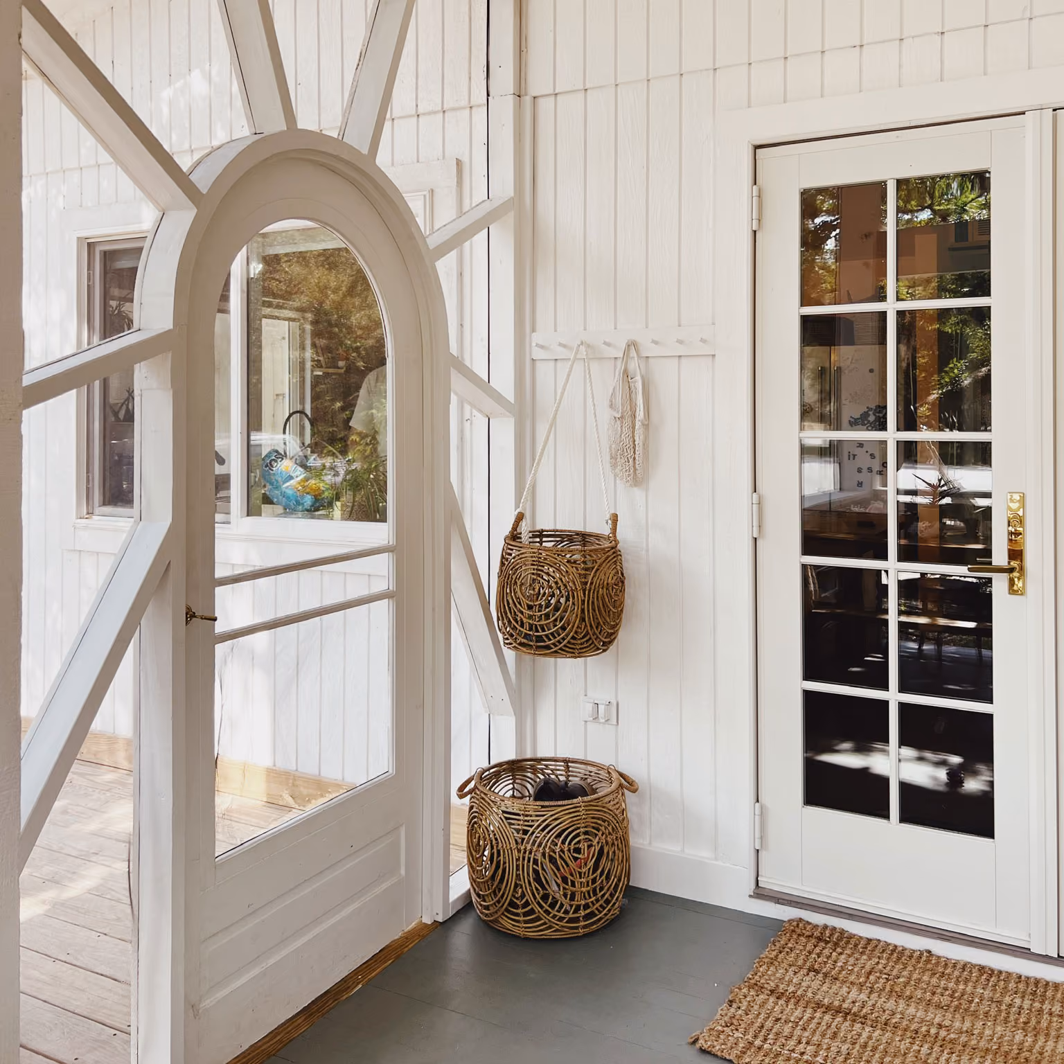 Bright entryway with a white arched screen door featuring sunray-style wooden beams, next to a glass-paneled white door with gold handle, two round woven baskets for storage—one hanging and one on the floor—and a textured natural fiber rug on a gray floor.