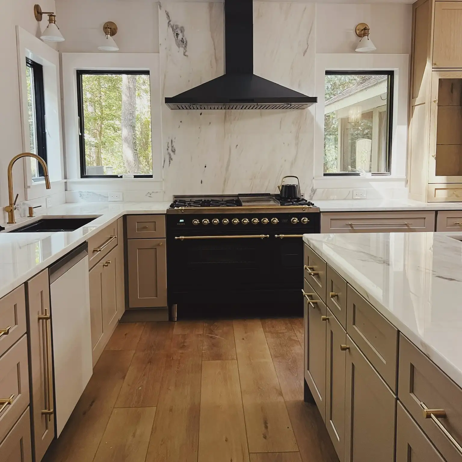 Modern kitchen with beige cabinets, white marble countertops, wood flooring, a black stove with brass knobs beneath a black range hood, two windows showing greenery outside, a brass faucet over a black sink, and brass cabinet handles.