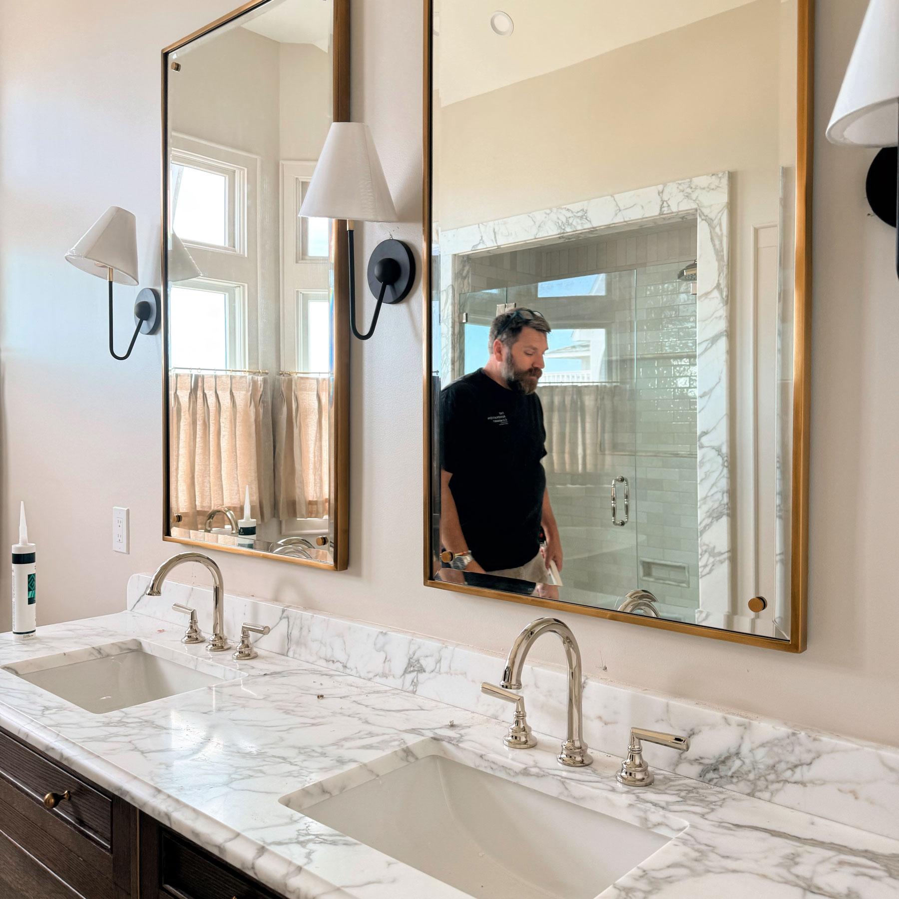 Modern bathroom countertop with white marble surface featuring two sinks and silver faucets, two rectangular mirrors with gold frames, and a man with a beard reflected in the right mirror standing near a shower area with glass doors and white subway tiles.