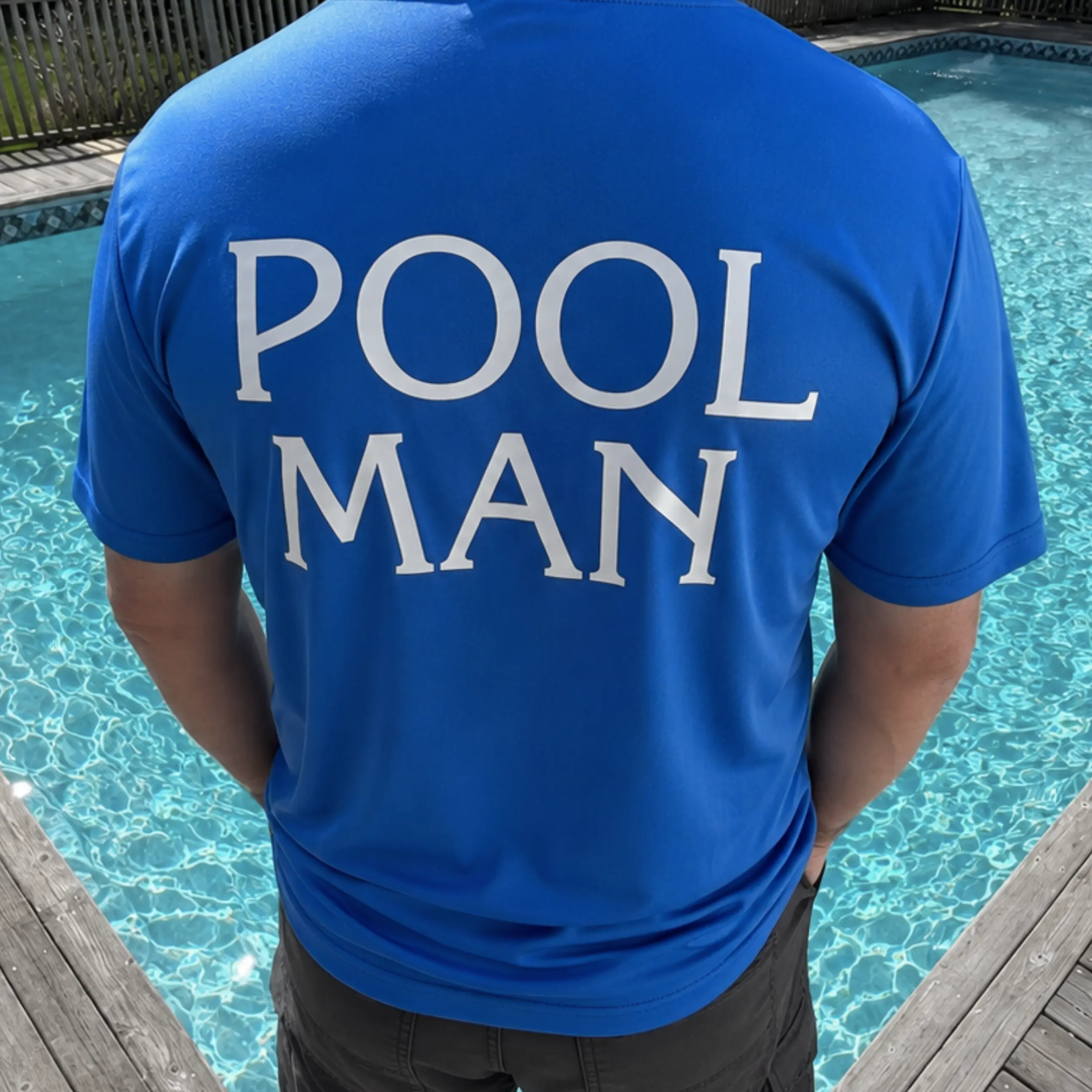 Person wearing a blue shirt with the words 'POOL MAN' printed in large white letters on the back, standing by a swimming pool with clear water and a wooden deck surrounding it.