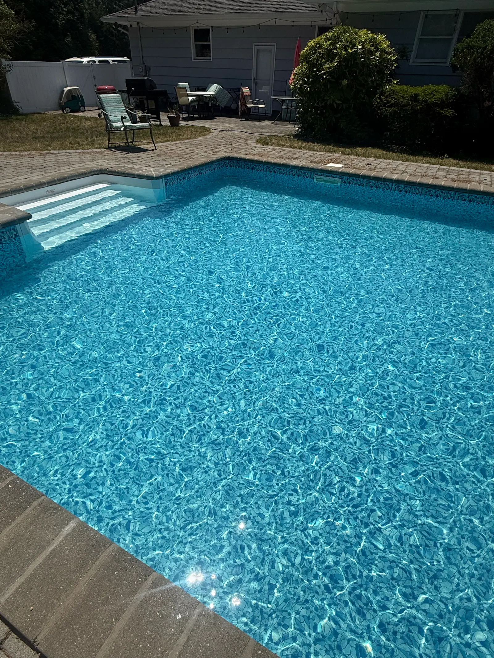 Clear blue swimming pool with sunlight reflecting on the water, steps leading into the pool, a stone tile edge, surrounded by lawn chairs, patio furniture, a grill, and a small playhouse near a grey house and bushes in Hampton Bays.