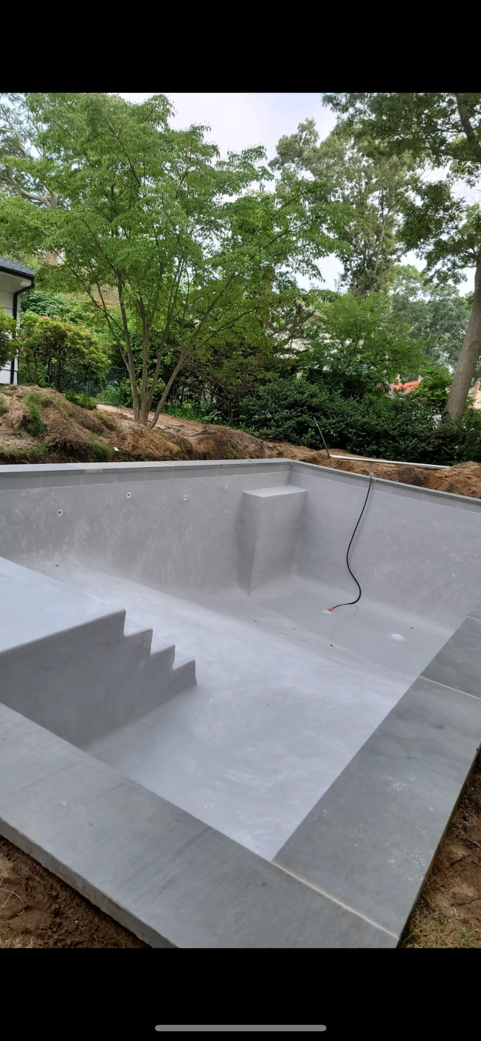 Empty gray concrete swimming pool under construction with steps on the left side, a raised platform on the far side, and a black cable hanging inside, surrounded by green trees and bushes in East Quogue.