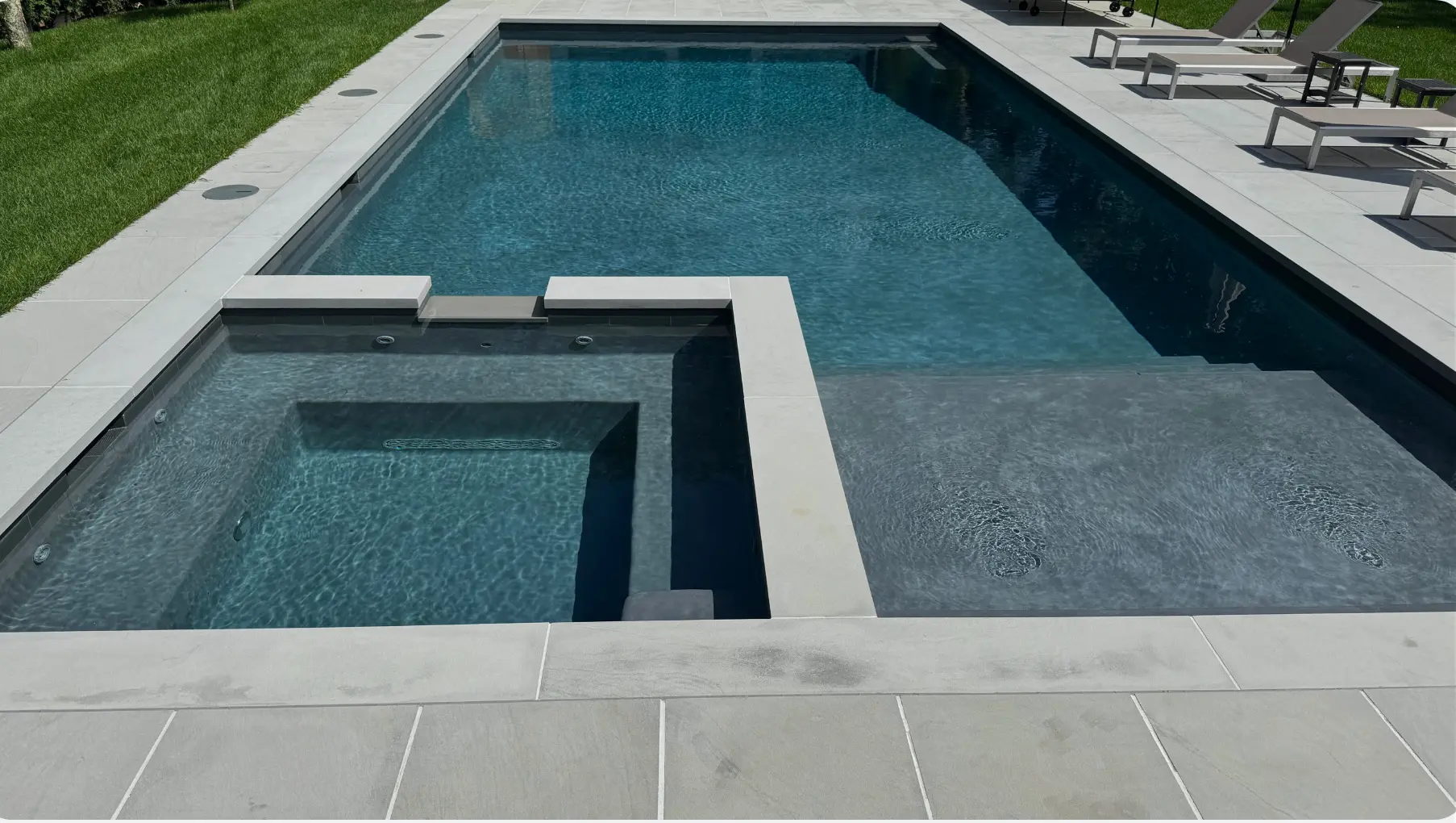 Modern backyard in Remsenburg with a rectangular swimming pool featuring a built-in hot tub on the left and a shallow tanning ledge on the right, surrounded by light stone decking and green lawn with chaise lounges in the background.