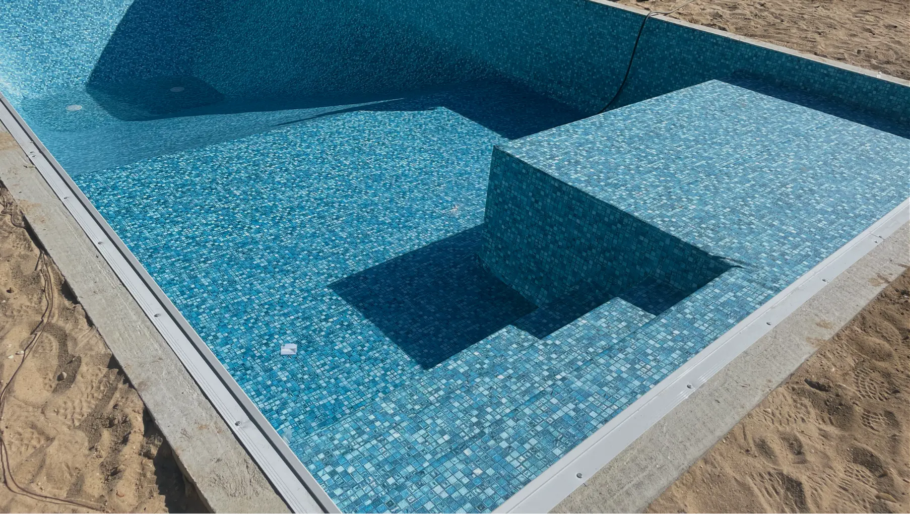 Empty rectangular swimming pool with blue mosaic tile interior, shallow steps on one side, and surrounding sandy ground in Westhampton.