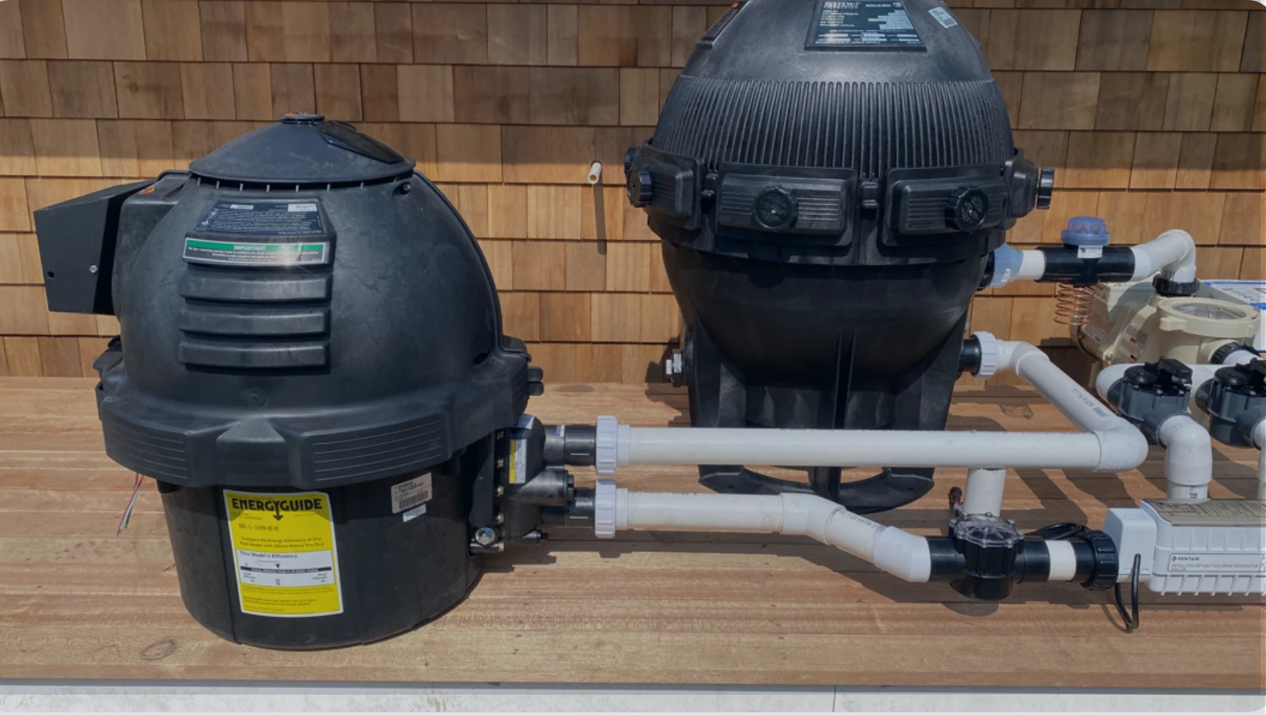 Two large black pool filtration units connected by white PVC pipes and valves on a wooden deck against a wooden shingle wall.