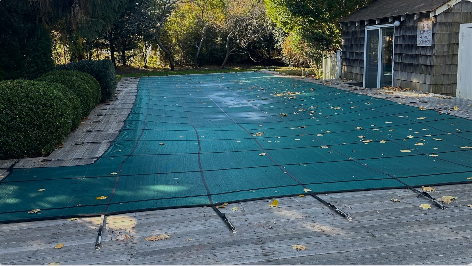 Outdoor swimming pool covered with a green safety cover secured by straps, surrounded by wooden decking, some fallen leaves scattered on the cover and deck, with bushes and trees in the background and a small wooden pool house to the right in East Quogue.