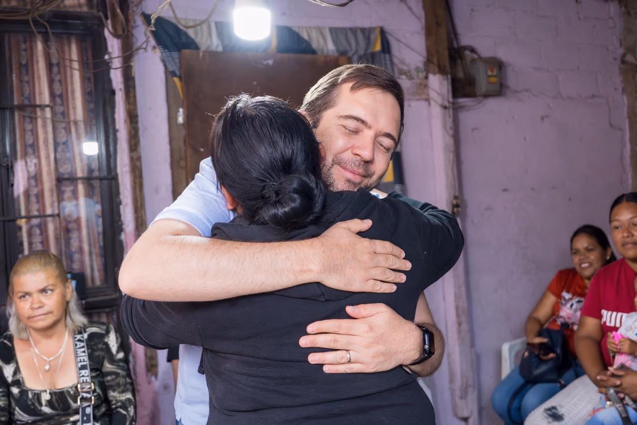 Man and woman warmly hugging indoors with three seated women watching in a modest room.