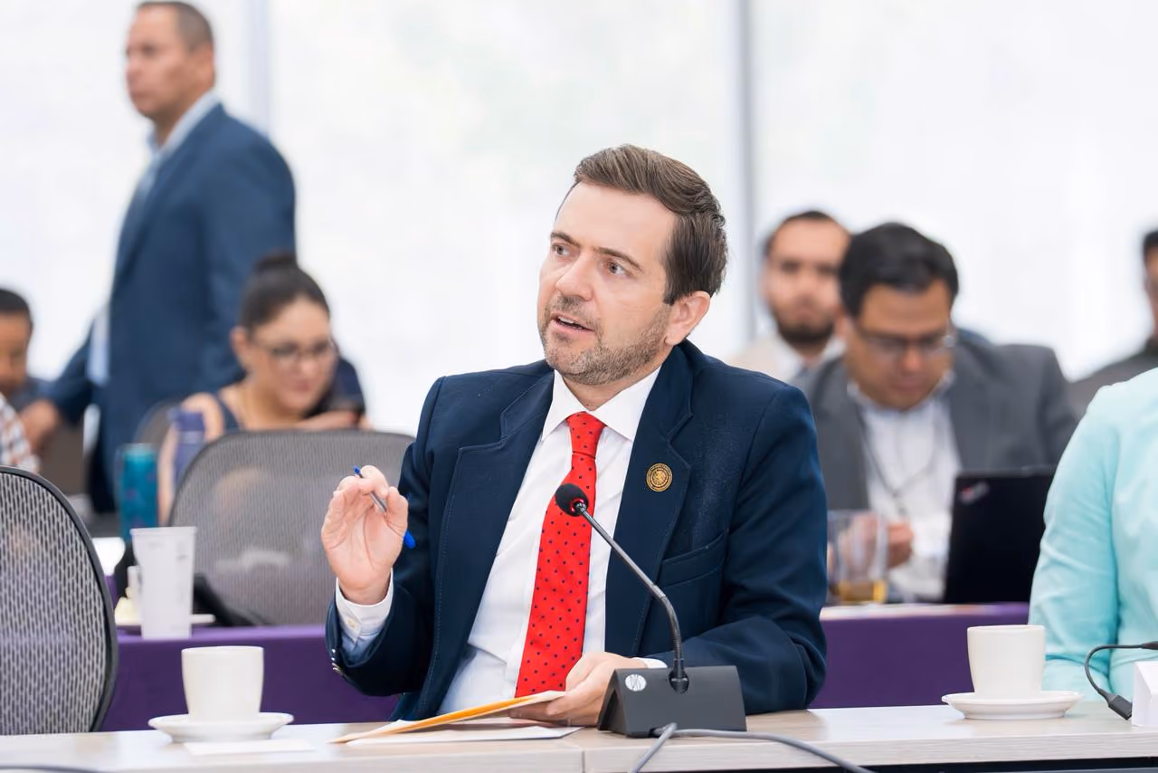 Man in suit and red tie speaking at a conference table with microphone and coffee cups.
