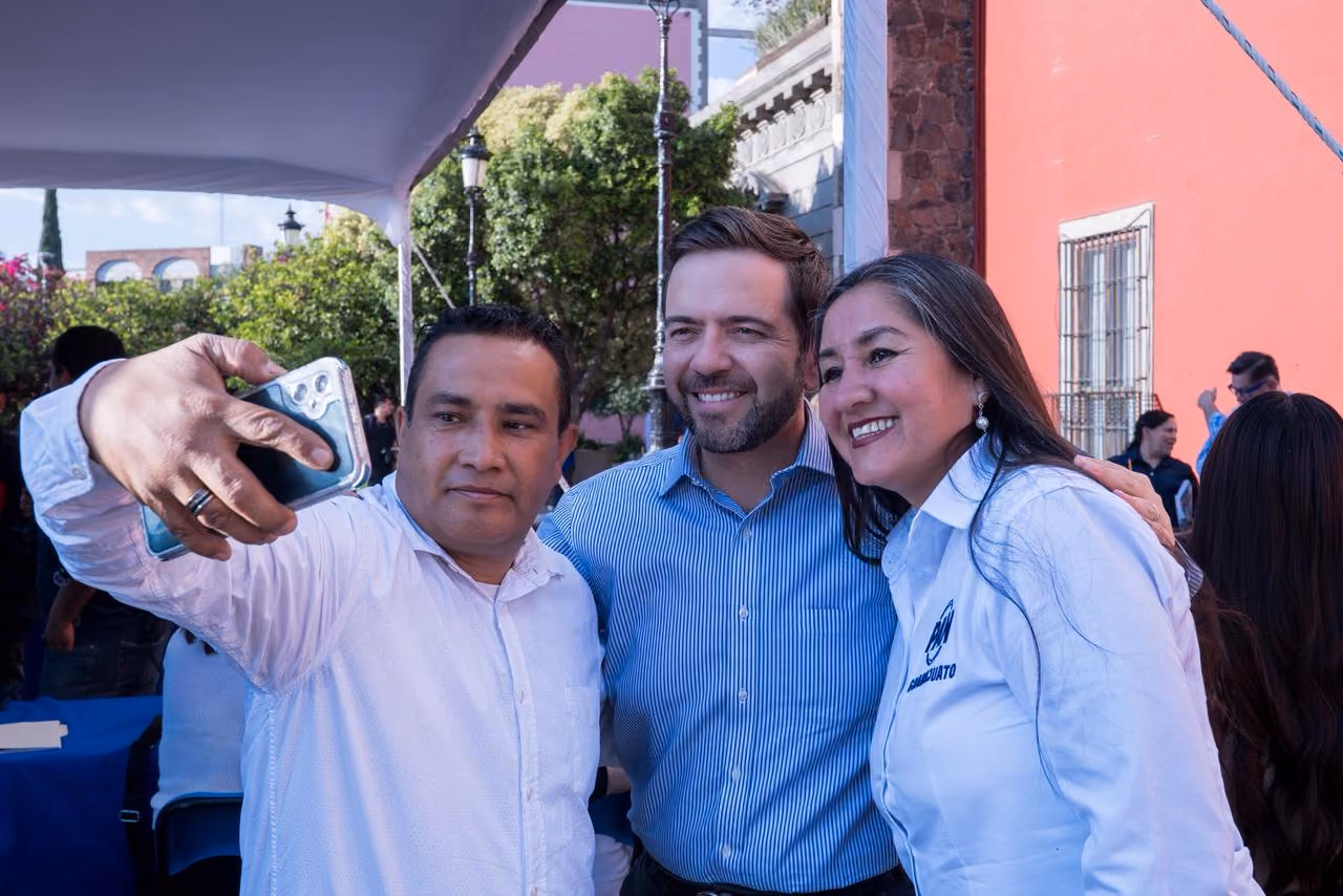 Three smiling adults pose closely for a selfie outdoors under a tent with trees and buildings behind.