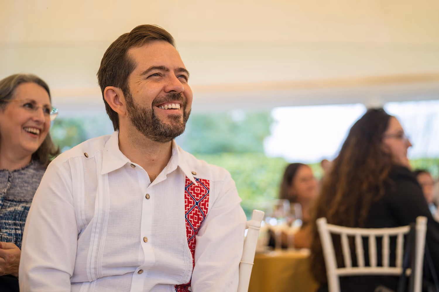 Smiling man with beard in white shirt with red pattern seated among other people at event.