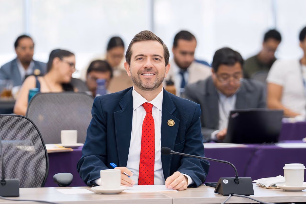 Man in a suit with a red tie smiling at a conference table with people working in the background.