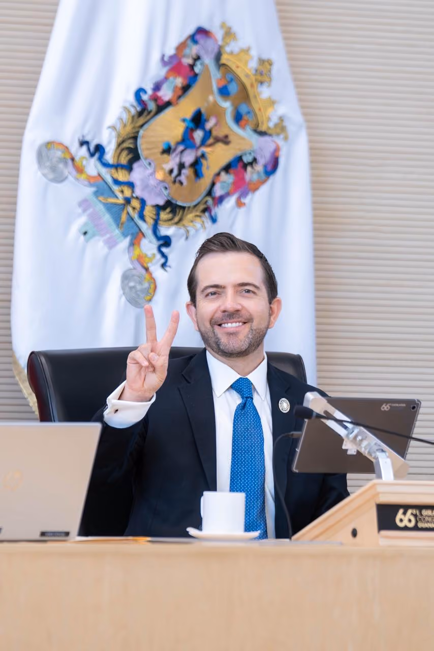 Smiling man in suit and blue tie shows peace sign seated at desk with emblem behind him.
