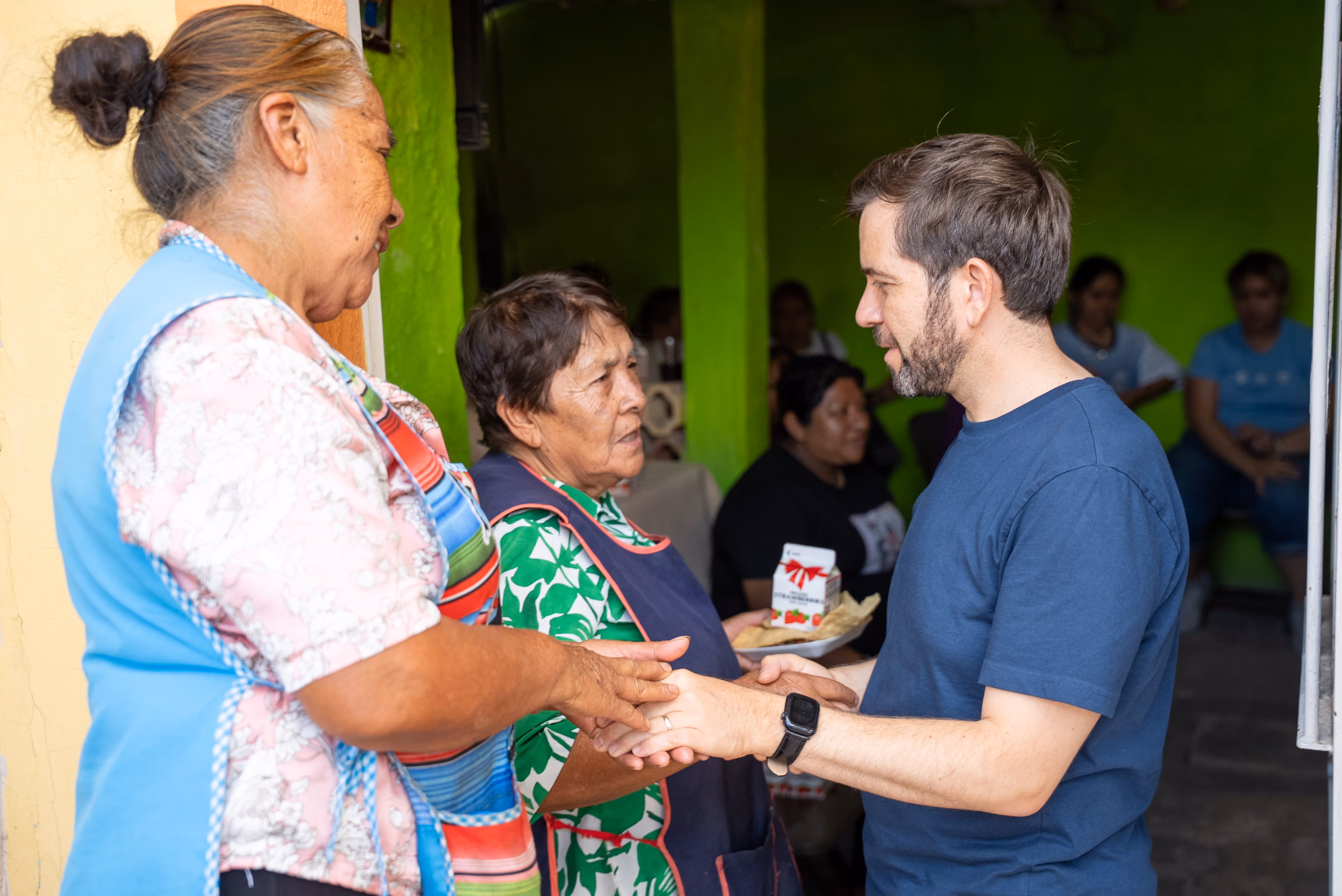 Man in blue shirt holds hands and talks with two older women wearing aprons indoors.