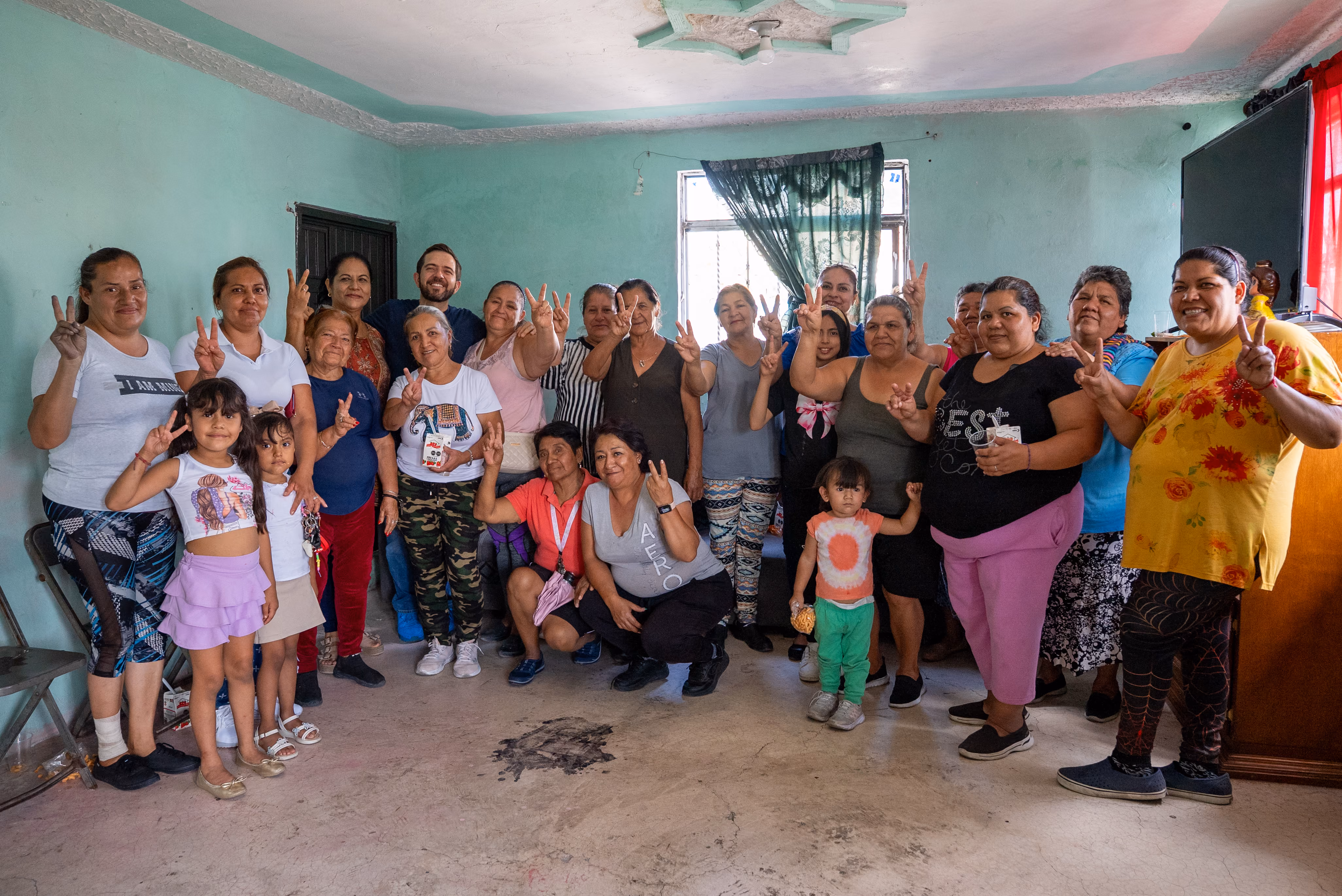 Group of diverse people, women and children, smiling and making peace signs indoors.